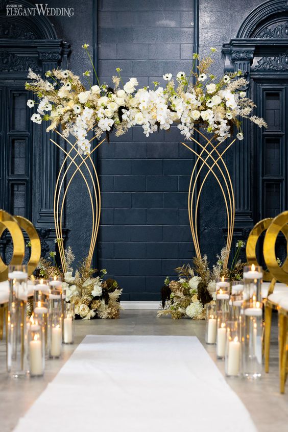 a sophisticated gold and white wedding ceremony space with a gold altar with white blooms, white and gold chairs and pillar candles in glasses