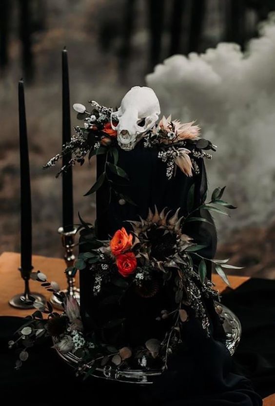 a statement black wedding cake with bright and white blooms, dried grasses and husks plus a skull for a Halloween wedding