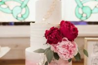 a white to pink ombre wedding cake decorated with gold leaf, with burgundy and pink blooms and greenery