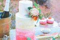 a white, yellow and pink watercolor and ombre wedding cake is topped with a flirty gold cake topper