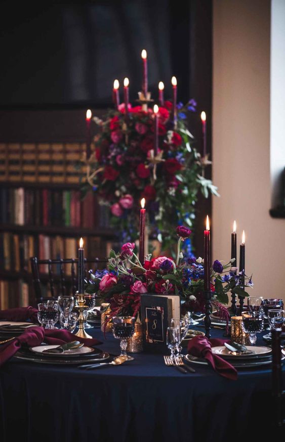 an exquisite Halloween wedding tablescape with a navy tablecloth, purple napkins, a lush centerpiece of purple, pink and burgundy blooms and bloody candles