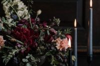 a beautiful, textural and sophisticated floral wedding centerpiece of burgundy and peachy blooms, greenery and thin and tall candles
