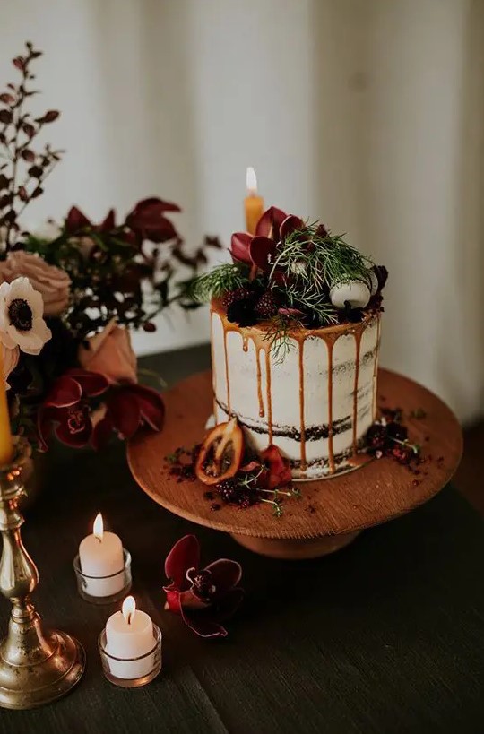 a chocolate naked wedding cake with caramel drip, fresh berries and dark blooms, macarons and greenery