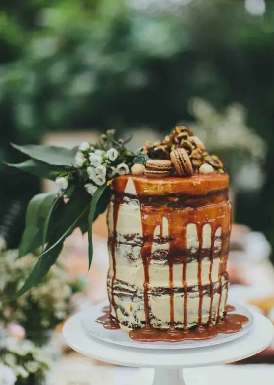 a chocolate naked wedding cake with caramel drip, macarons, nuts, greenery and white blooms is amazing