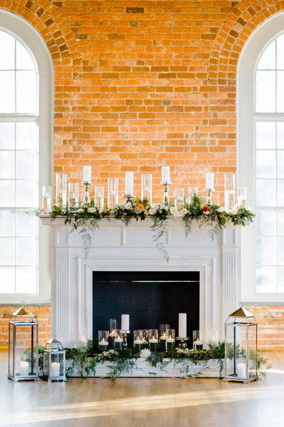 a faux fireplace decorated with greenery and bold blooms, lots of candles in candle holders and lanterns