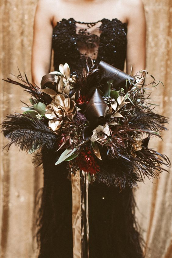 a glam Halloween wedding bouquet with red and gilded blooms and leaves, greenery, dark feathers and gold ribbons