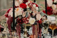 a gorgeous decadent Halloween wedding centerpiece of a tall gold bowl with blush and deep red blooms, greenery, grapes and with black candles around