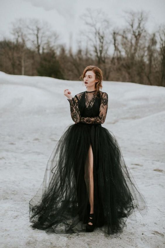a modern black wedding dress with a lace top, a tulle skirt with a front slit and long sleeves, black shoes and a moody lipstick