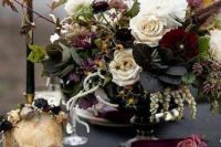 a moody and refined Halloween wedding centerpiec eof neutral and burgundy blooms, greenery and dark foliage, black candles and skulls
