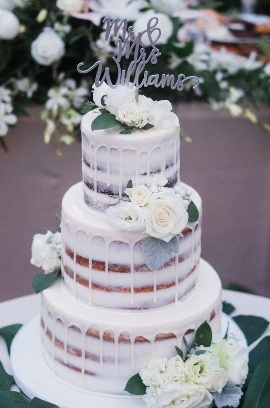 a naked three-tier wedding cake with creamy drip, white blooms and leaves and a calligraphy topper is a delicious option