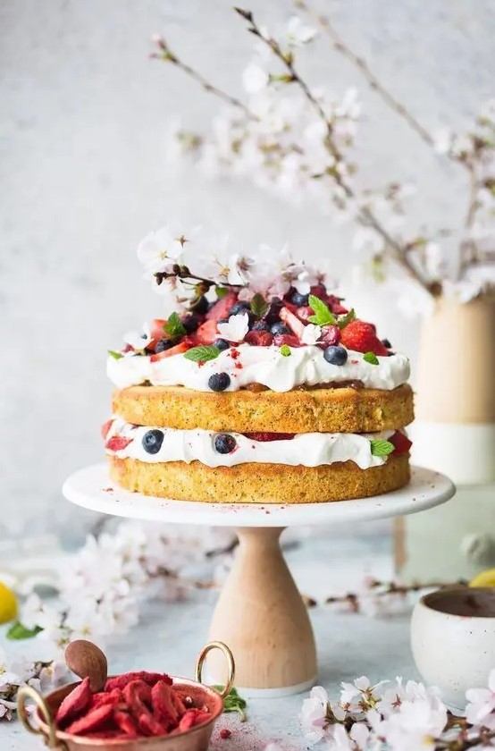 a naked wedding cake topped with fresh whipped cream and berries and cherry blossom