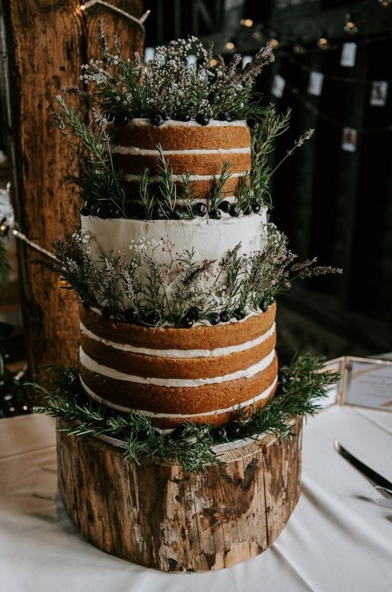 a partly naked wedding cake with lots of greenery, wildflowers and fresh berries for a rustic wedding
