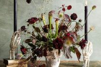 a scary and chic Halloween wedding centerpiece of a skull as a vase, dark and red blooms, black candles holded by skeleton hands is a lovely idea