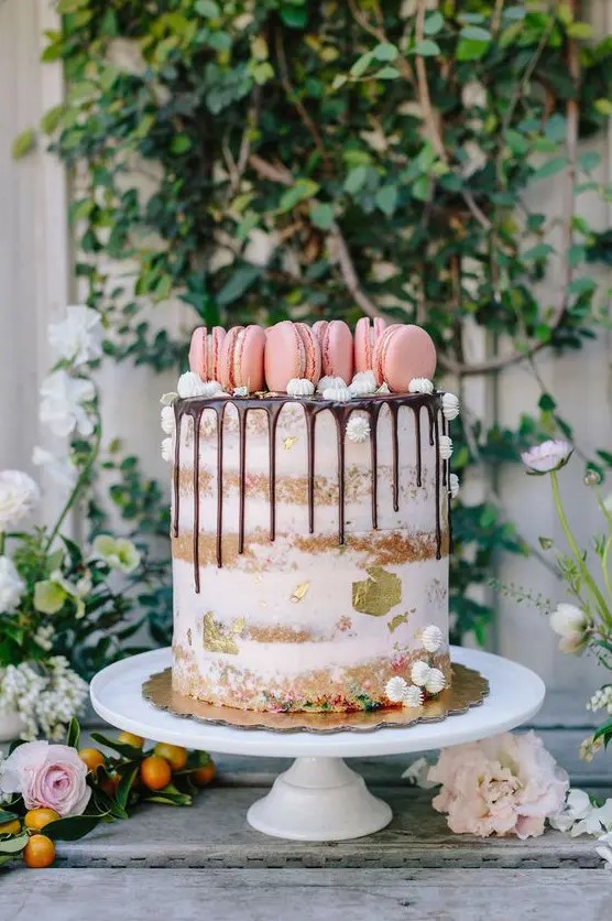 a semi naked cake with gold foil and topped with pink macarons on top plus small meringues
