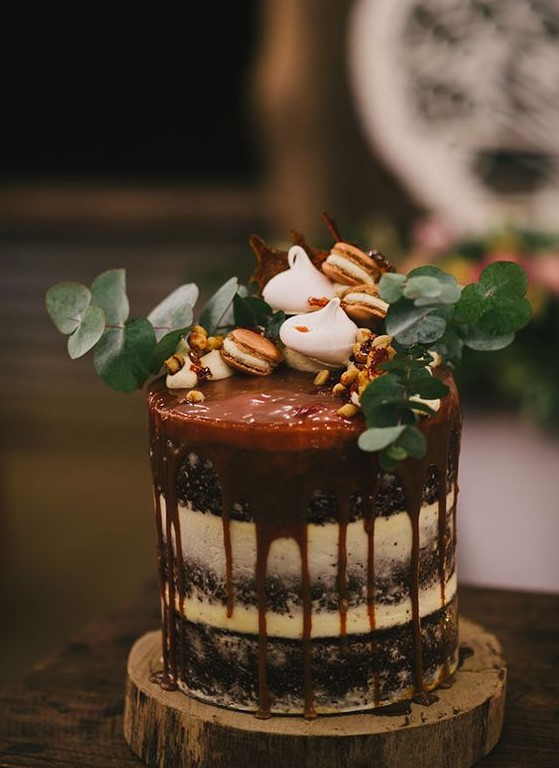 a semi naked wedding cake with caramel drip, topped with eucalyptus, macarons and meringues