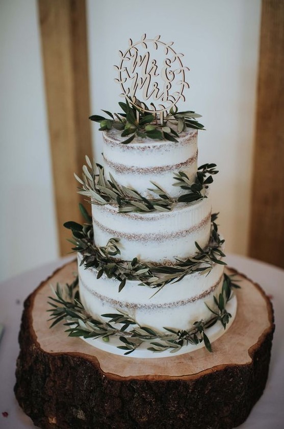 a simple naked three-tier wedding cake with fresh greenery and olives and a fun carved topper is a lovely idea