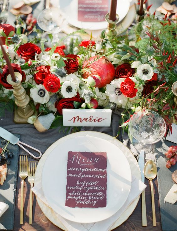 a bold Christmas wedding centerpiece of red and white blooms, pomegranates, greenery and burgundy candles in gold candleholders