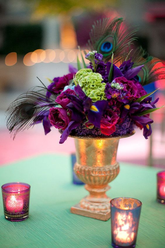 a bold wedding centerpiece with a gold urn, deep purple blooms and green ones plus peacock feathers