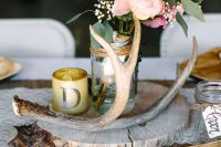 a bright rustic wedding centerpiece of a wood slice, antlers, a candle in a glass and bright blooms in a jar