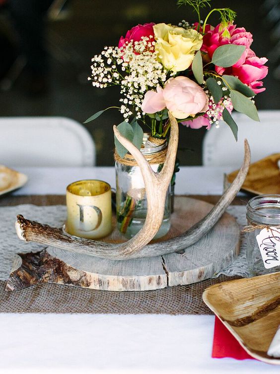 a bright rustic wedding centerpiece of a wood slice, antlers, a candle in a glass and bright blooms in a jar