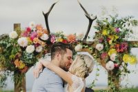 a bright wedding arch decorated with bright blooms, greenery and with antlers on top is a bold and chic idea