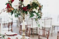 a chic Christmas tablescape with a greenery and red bloom runner and a matching tall centerpiece, burgundy napkins and gold chargers