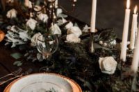 a chic and refined Christmas tablescape with an evergreen runner with white roses, tall and thin candles, gold chargers and white porcelain