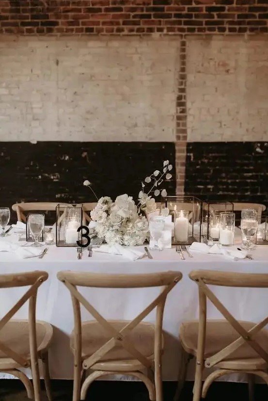 a chic winter wedding tablescape done in white, with candle lanterns, a lush white floral centerpiece, a black table number