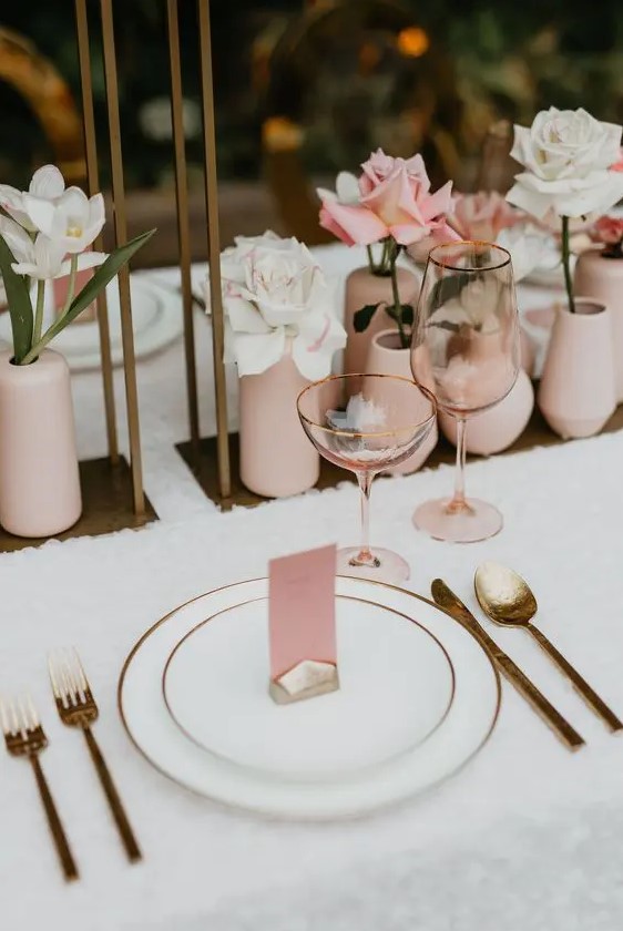 a lovely modern wedding tablescape with a neutral tablecloth, blush vases with a single bloom in each, gold-rimmed plates and gold cutlery