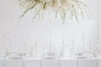 a minimalist ethereal wedding reception space with an oversized overhead floral decoration, sheer chairs and everything white
