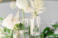 a modern neutral wedding centerpiece of greenery, white blooms and dried leaves in clear vases and small candles in glasses