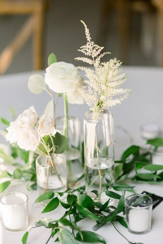 a modern neutral wedding centerpiece of greenery, white blooms and dried leaves in clear vases and small candles in glasses