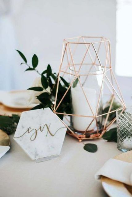 a modern wedding centerpiece of a copper lantern with candles, greenery, smaller candles, a marble table number is a lovely idea