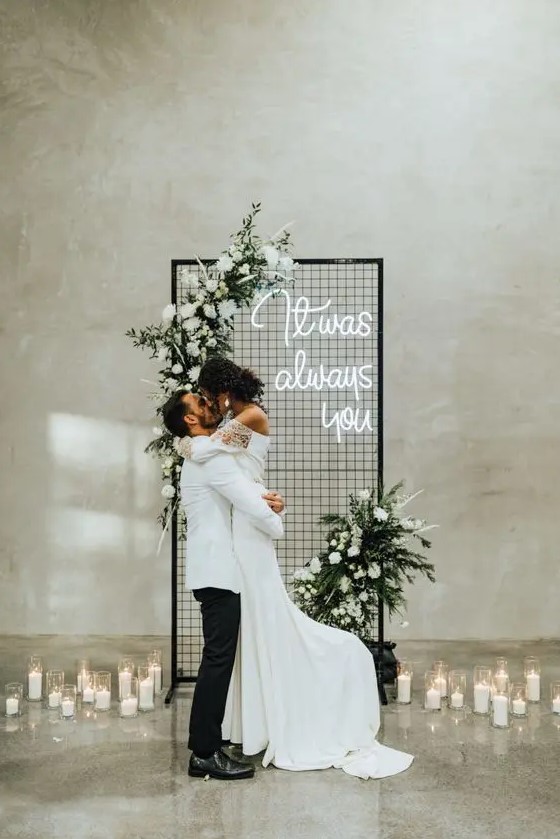 a modern winter wedding backdrop with white blooms and greenery, a neon sign and pillar candles around