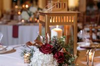 a refined Christmas wedding centerpiece of burgundy and white blooms, pinecones, greenery and a gold candle lantern is chic