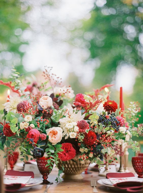 a refined holiday wedding centerpiece of a gold bowl, blush, red and burgundy blooms, greenery, berries and red candles around