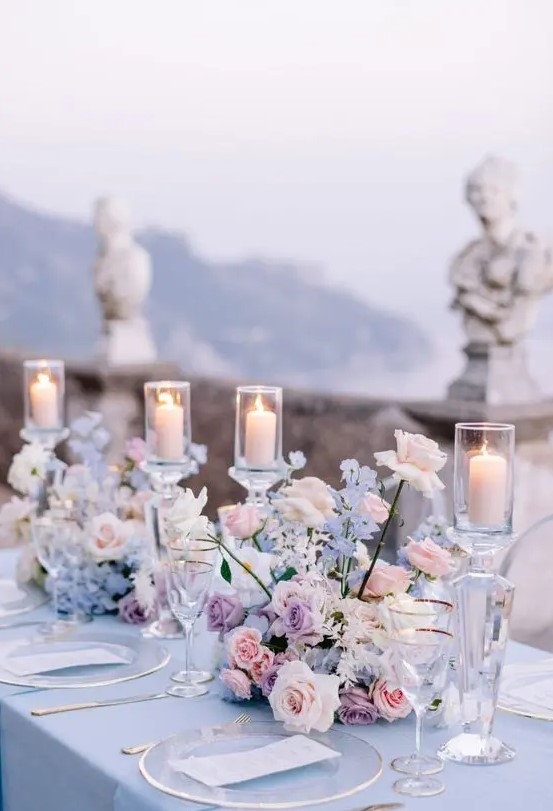 a refined wedding tablescape with a blue tablecloth, blush, blue and lilac blooms, candles and clear chargers and glasses