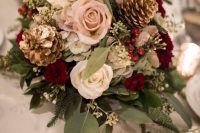 a romantic Christmas wedding centerpiece of burgundy, blush, white blooms, greenery, gilded pinecones and candles around