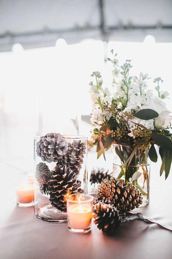 a rustic Christmas wedding centerpiece of glass vases with pinecones, a candle and some white blooms and greenery