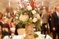 a rustic Christmas wedding centerpiece of white blooms, thistles, berries, twigs and greenery and a vase wrapped with bark