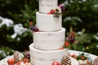a semi naked Christmas wedding cake topped with dark blooms, berries, apples and gilded pinecones