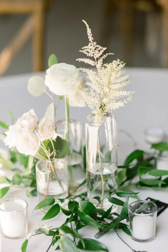 a subtle cluster wedding centerpiece of clear vases, candles, white ranunculus, leaves and roses plus greenery on the table is amazing