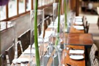 a very laconic modern wedding tablescape with an uncovered table, tall glass vases with allium is a chic and lovely idea