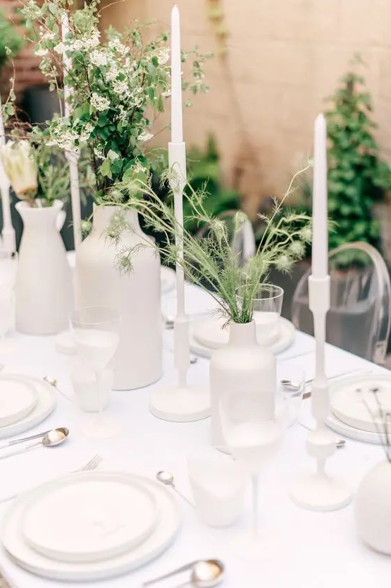 a very simple and minimal wedding tablescape with all white everything, with greenery and white blooms, tall and thin white candles and some cutlery