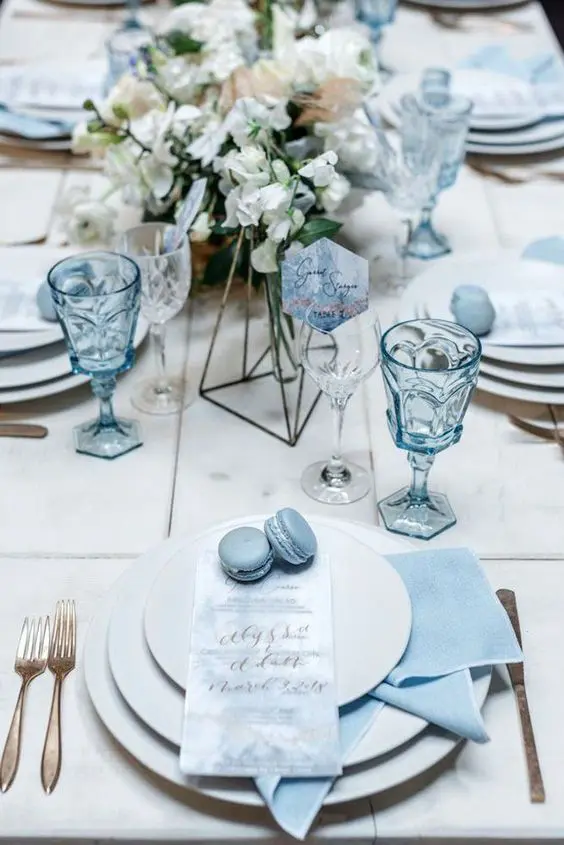 a white and ice blue winter wedding tablescape with blue napkins, glasses and macarons plus gold cutlery is chic