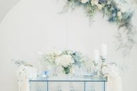 a white backdrop decorated with white and ice blue blooms and an ice blue sheer glass table and blooms for an ethereal look