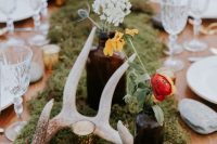 a woodland wedding table with a moss runner, candles, bright blooms in apothecary bottles and antlers is very chic