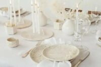 an airy white winter wedding tablescape with speckled plates, tall and thin white candles, dried branches with leaves and neutral linens