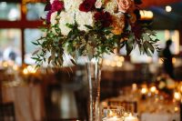 an exquisite Christmas wedding centerpiece of a tall glass vase, greenery, white, burgundy and blush blooms and lots of candles around
