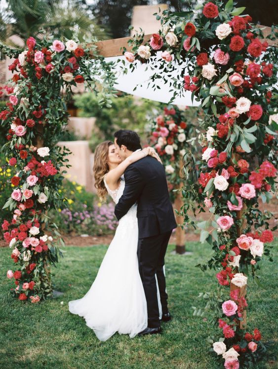 a bold wedding arch of red, blush and white blooms and greenery and sheer fabric is a chic idea